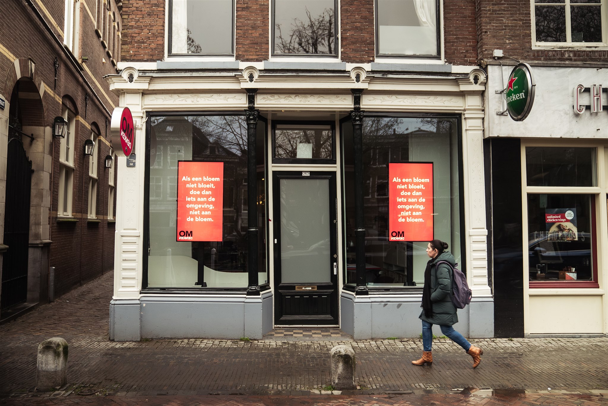 Marketing in Beeld Een persoon met een rugzak loopt langs een bakstenen gebouw met grote ramen waarop rode posters met Nederlandse tekst hangen. De straat is leeg, behalve de voetganger, en boven een deuropening rechts hangt een bordje van Heineken.