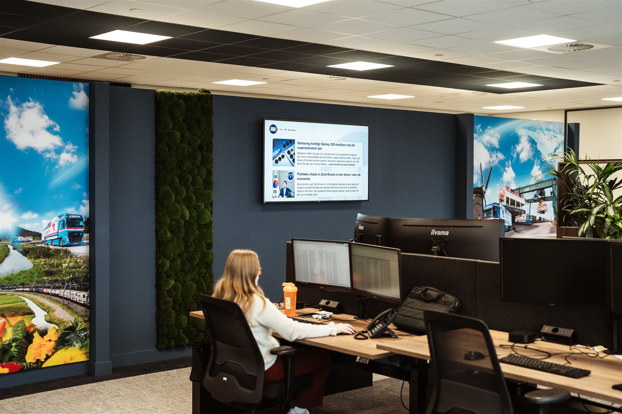 Marketing in Beeld Een persoon zit aan een bureau met meerdere monitoren in een modern kantoor. Grote muurschilderingen van buitenscènes en een groen mospaneel sieren de ruimte. Een scherm aan de muur toont bedrijfsnieuws of updates.
